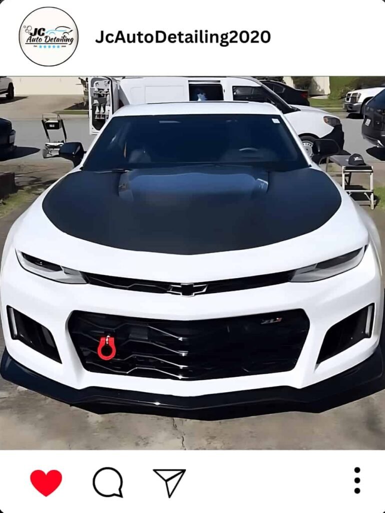 A white Chevrolet Camaro ZL1 with a black hood, recently detailed, parked in a parking lot. The car has a red tow hook and aggressive front styling